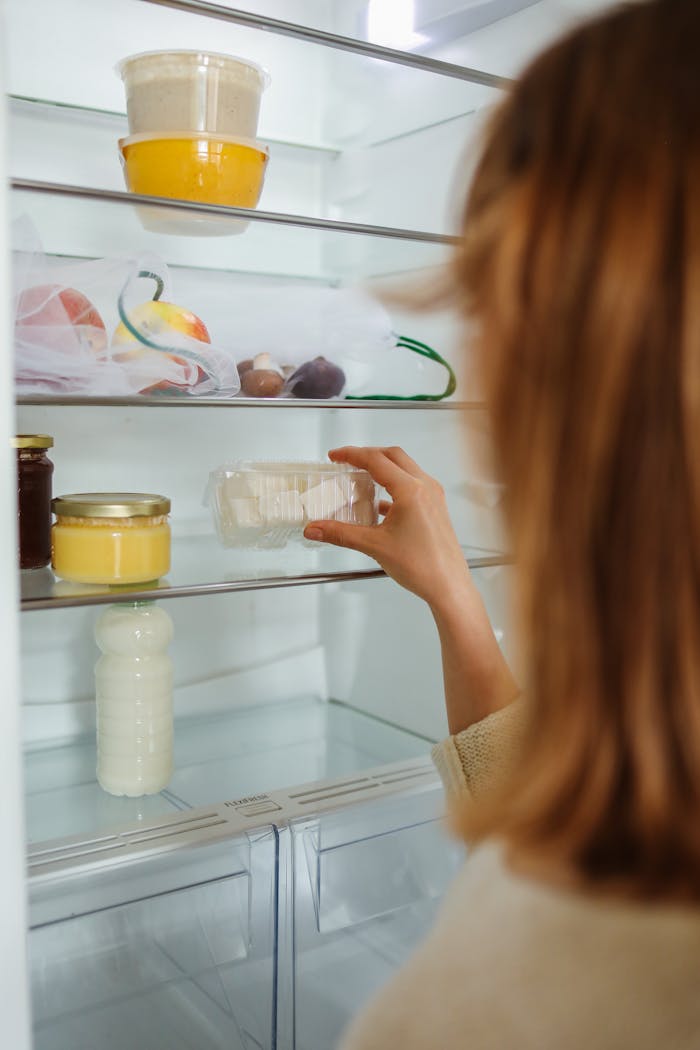 Woman organizing fridge shelves with assorted food containers. Close-up of hand reaching for a jar.